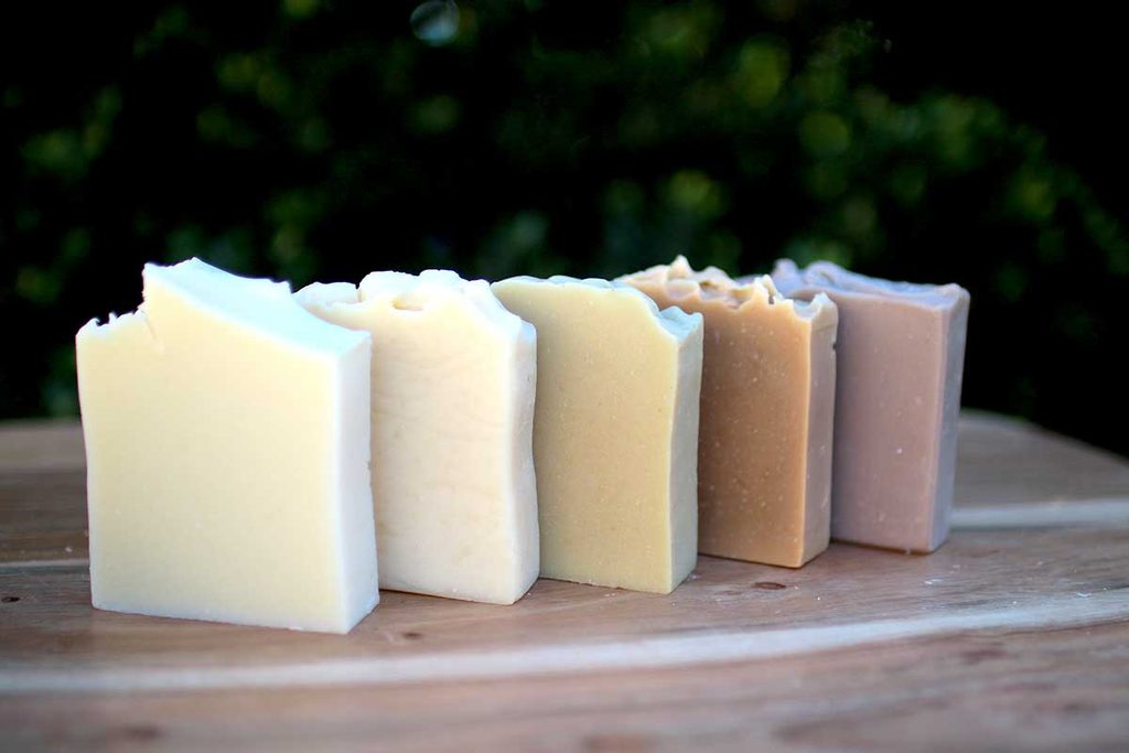 Five bars of handmade soap in shades of cream, beige, brown, and lavender, with textured tops, arranged on a wooden surface outdoors